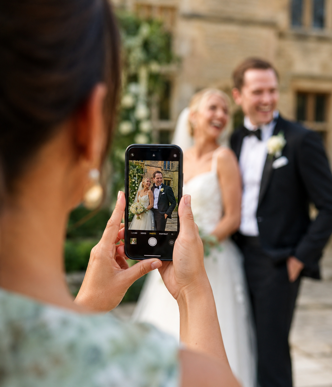 An over-the-shoulder view of a wedding guest using a smartphone to photograph the newly married couple at a UK wedding reception.
